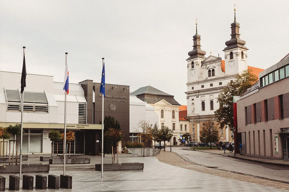 Площадь перед современным корпусом с надписью Trnavská univerzita и флагами, справа барочный собор с двумя башнями