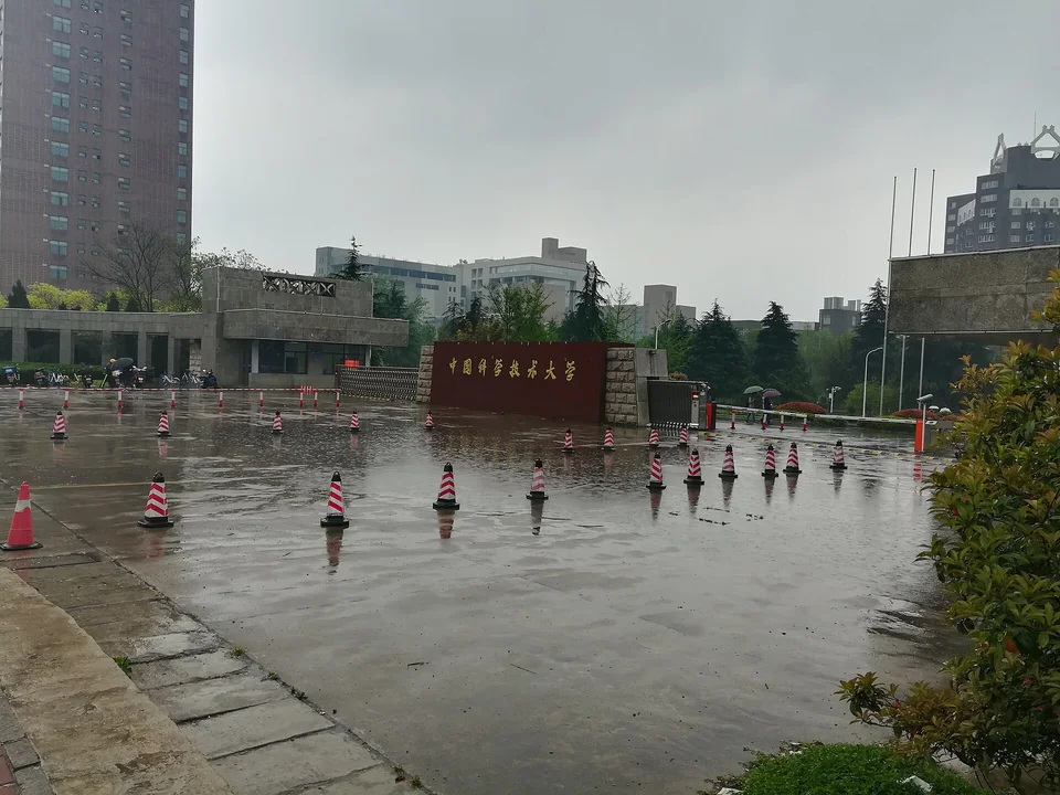 Мокрая после дождя площадь с красно-белыми конусами перед бордовой стеной с золотой надписью «中国科学技术大学», за ней корпуса кампуса и деревья
