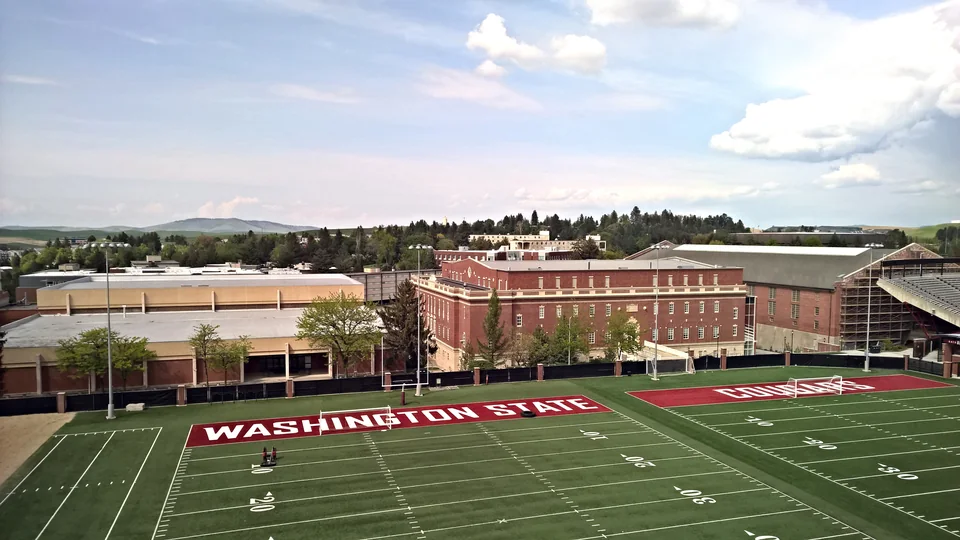 Зелёное футбольное поле с надписью Washington State, за ним корпуса из красного кирпича и холмы на горизонте