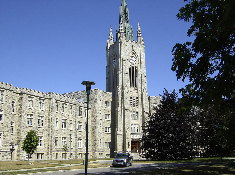 Grey-stone Collegiate Gothic building with a tower, a road and trees in front